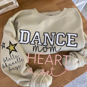 May include: Tan sweatshirt with the words "DANCE mom" in black and white block letters. The left sleeve has a gold glitter star and the names "stella, shanelle, sage" in black script. The word "HEART set" is in pink script.