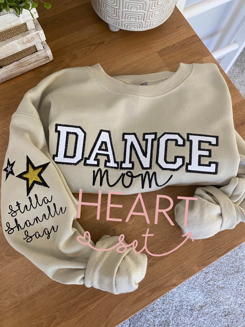 May include: Tan sweatshirt with the words "DANCE mom" in black and white block letters. The left sleeve has a gold glitter star and the names "stella, shanelle, sage" in black script. The word "HEART set" is in pink script.