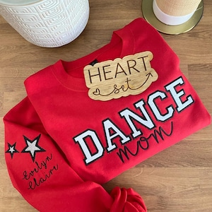 May include: A red sweatshirt with the words "DANCE mom" in white and black lettering. The sleeve has two star graphics and the name "evlyn claire" in cursive. A wooden plaque that says "HEART set" is on top of the sweatshirt.