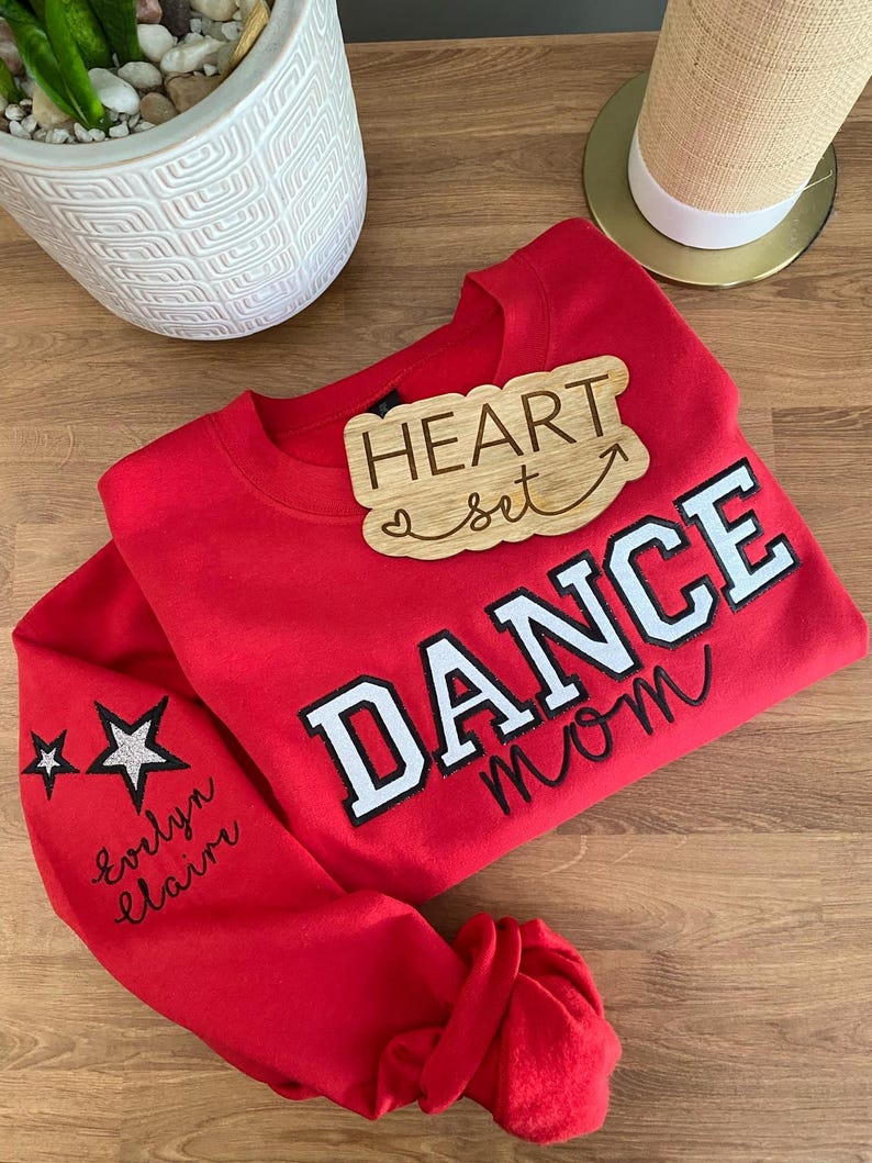 May include: A red sweatshirt with the words "DANCE mom" in white and black lettering. The sleeve has two star graphics and the name "evlyn claire" in cursive. A wooden plaque that says "HEART set" is on top of the sweatshirt.