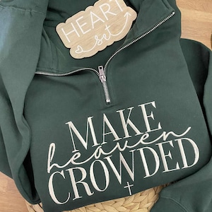 May include: Dark green quarter-zip sweatshirt with the text "MAKE heaven CROWDED" in white script. A small wooden sign that says "HEART set" is placed on the sweatshirt. The sweatshirt is folded on a woven mat.