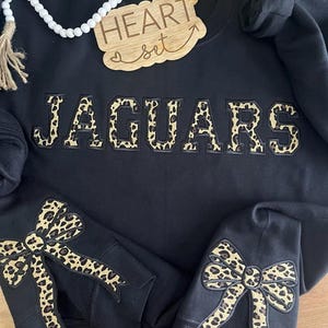 May include: Black sweatshirt with the word "JAGUARS" in leopard print letters. The sweatshirt has leopard print bows on the sleeves and a wooden sign that says "HEART set" above the word "JAGUARS".