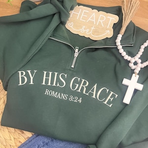 May include: A dark green pullover sweatshirt with the text "BY HIS GRACE ROMANS 3:24" in white. The sweatshirt has a silver zipper and is paired with blue jeans. A wooden sign that says "HEART set" and a white cross necklace are also in the image.
