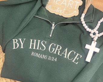 May include: A dark green pullover sweatshirt with the text "BY HIS GRACE ROMANS 3:24" in white. The sweatshirt has a silver zipper and is paired with blue jeans. A wooden sign that says "HEART set" and a white cross necklace are also in the image.