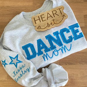 May include: A light gray sweatshirt with the words "DANCE mom" in blue, glittery letters. The sleeve has two blue stars and the name "Tessa Presley" in cursive. A wooden sign that says "HEART set" is on top.