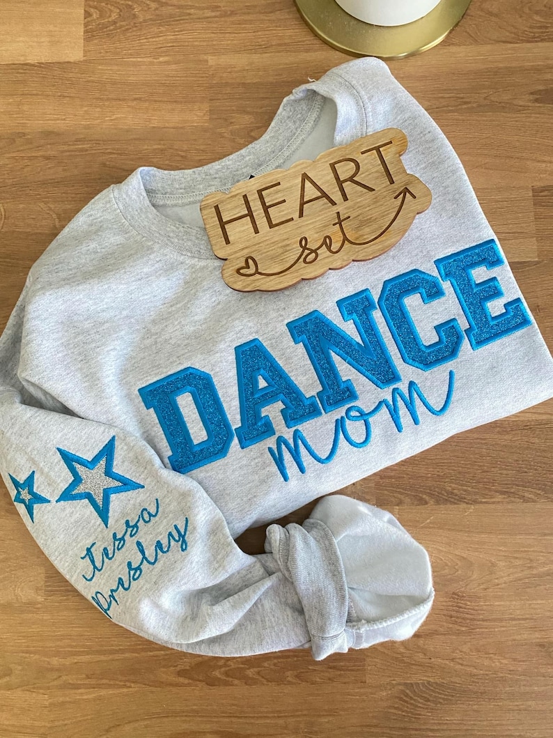 May include: A light gray sweatshirt with the words "DANCE mom" in blue, glittery letters. The sleeve has two blue stars and the name "Tessa Presley" in cursive. A wooden sign that says "HEART set" is on top.