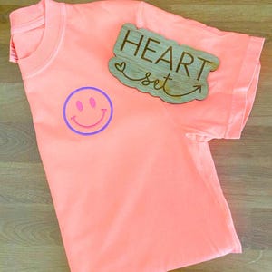 Op de afbeelding: Een koraalkleurig t-shirt met een paarse en roze smiley-gezichtsgrafiek. Een houten bord met de woorden "HEART set" en een pijl is op het shirt geplaatst. Het t-shirt is opgevouwen en toont de kraag en de mouw.
