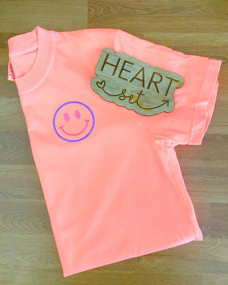 Op de afbeelding: Een koraalkleurig t-shirt met een paarse en roze smiley-gezichtsgrafiek. Een houten bord met de woorden "HEART set" en een pijl is op het shirt geplaatst. Het t-shirt is opgevouwen en toont de kraag en de mouw.