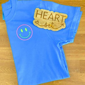 Op de afbeelding: Een lichtblauw t-shirt met een smiley-gezicht en de tekst "HEART set" op een houten uitsparing. De smiley heeft een roze omtrek en groene ogen en mond. Het shirt is op een houten oppervlak gevouwen.