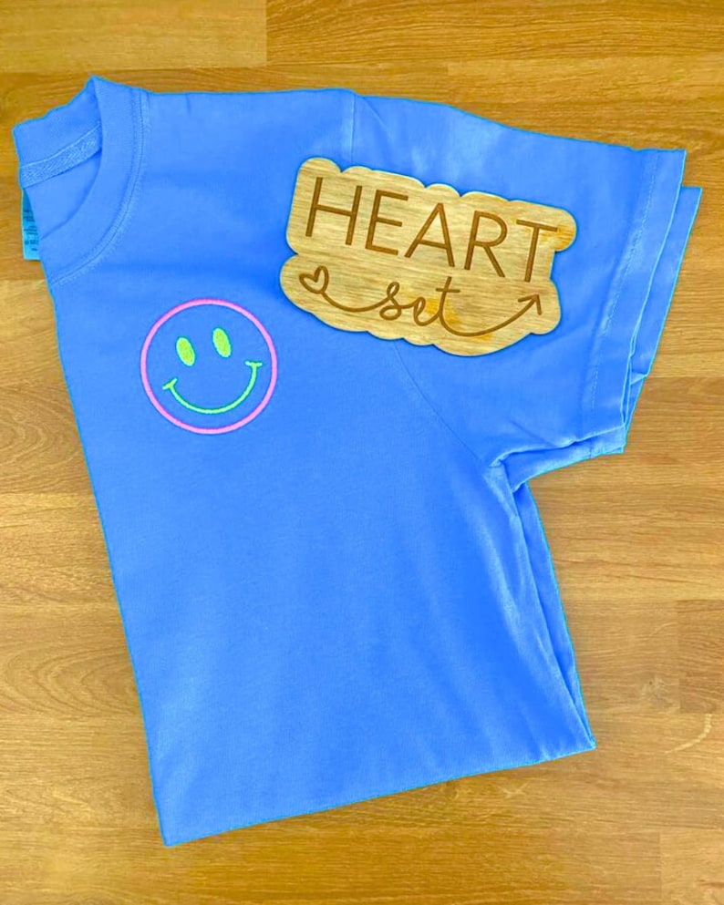 Op de afbeelding: Een lichtblauw t-shirt met een smiley-gezicht en de tekst "HEART set" op een houten uitsparing. De smiley heeft een roze omtrek en groene ogen en mond. Het shirt is op een houten oppervlak gevouwen.