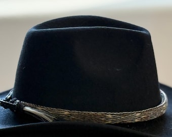 May include: A black felt fedora hat featuring a structured crown and a wide brim. The base of the crown is encircled by a decorative band with a woven geometric pattern in shades of brown and grey, finished with a small black side buckle.