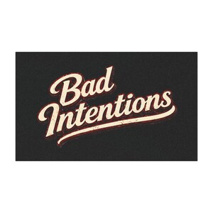 以下が含まれることがあります： ヴィンテージ風の「Bad Intentions」の文字がプリントされた、長方形の黒いドアマット。 文字はクリーム色で赤い縁取りがあり、レトロな雰囲気。 マットは質感のある外観です。