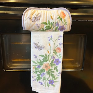 May include: A white kitchen towel with a floral pattern featuring butterflies, lavender, and pink flowers. The towel is hanging on an oven door handle.