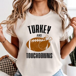 May include: A cream-coloured t-shirt featuring the text "TURKEY AND TOUCHDOWNS" in black. A brown American football and turkey leg graphic is below the word "AND". The shirt has short sleeves.
