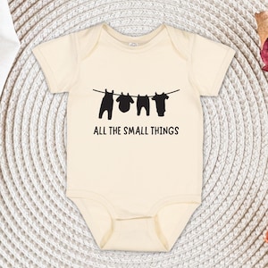 May include: Cream-colored baby onesie with a black graphic of clothes hanging on a clothesline. The text "ALL THE SMALL THINGS" is printed below the graphic. The onesie is made of soft material.