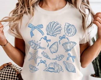 May include: Cream-colored t-shirt featuring a collection of blue seashell illustrations and bows. The design includes various shell shapes and a starfish, creating a coastal theme. The shirt is a casual, short-sleeved style.
