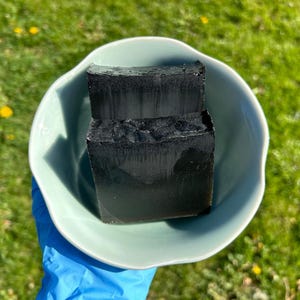 May include: Two black charcoal soap bars in a light blue ceramic bowl. The soap bars are stacked on top of each other.