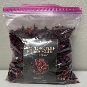 Peut inclure: Un sac en plastique transparent rempli de roselle d'hibiscus séchée. Le sac est fermé par une fermeture éclair rose et porte une étiquette indiquant "100% ORGANIC DRIED HIBISCUS ROSELLE (Hibiscus Sabdariffa)". Poids net : 454g.