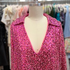 Puede incluir: Una blusa rosa de lentejuelas con cuello en V profundo y mangas largas. La blusa tiene un cuello y est&aacute; hecha de una tela brillante.