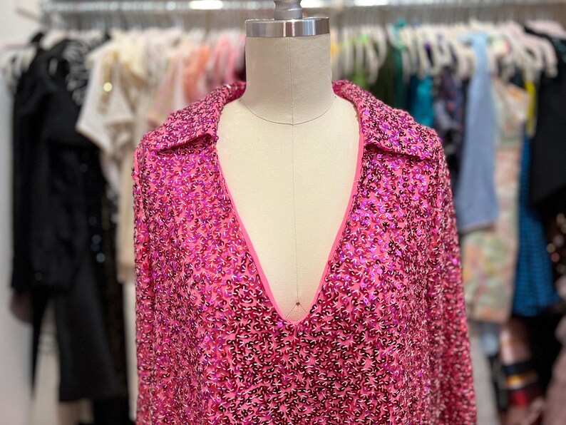 Puede incluir: Una blusa rosa de lentejuelas con cuello en V profundo y mangas largas. La blusa tiene un cuello y est&aacute; hecha de una tela brillante.