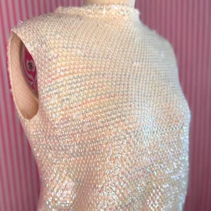 Op de afbeelding: Een mouwloze top volledig bedekt met iriserende, lichtreflecterende pailletten die een glinsterend, veelkleurig effect creëren op een crèmekleurige basis. Het kledingstuk is voorzien van een hoge, ronde halslijn en een getailleerd model.