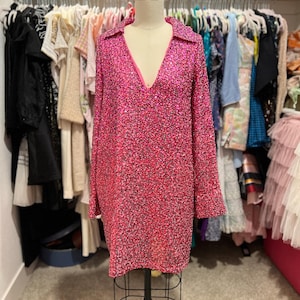 Puede incluir: Un vestido rosa de lentejuelas corto con mangas largas y escote en V. El vestido est&aacute; en un maniqu&iacute; en un armario.