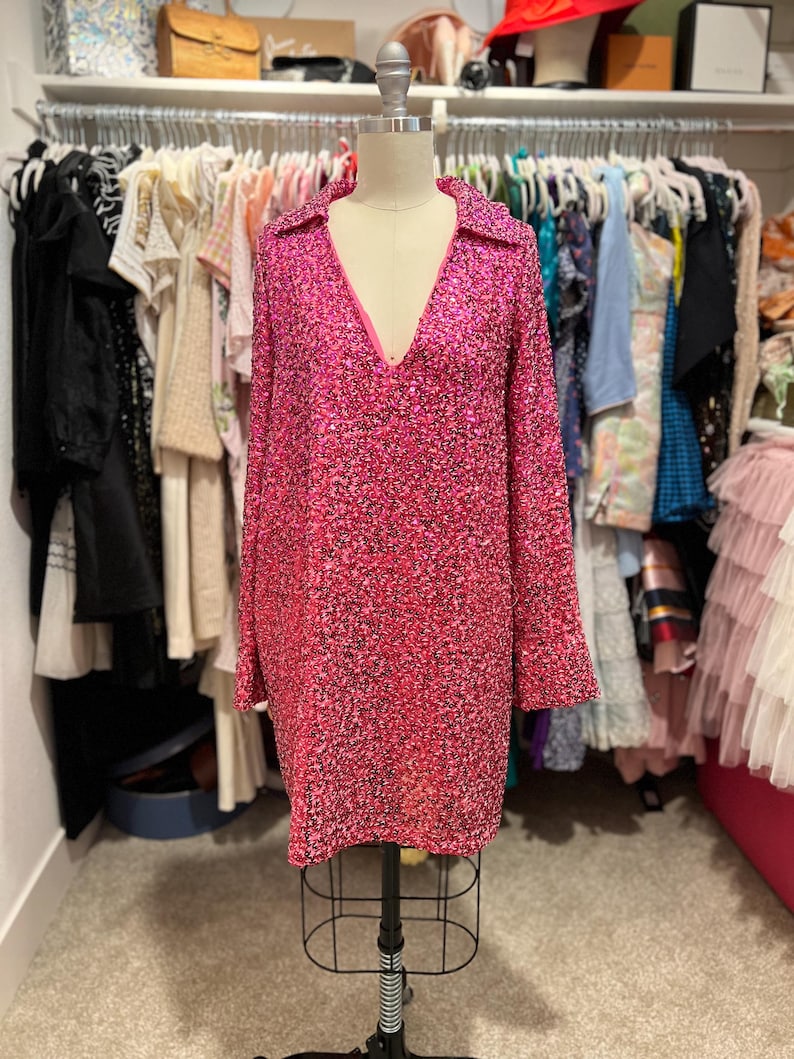 Puede incluir: Un vestido rosa de lentejuelas corto con mangas largas y escote en V. El vestido est&aacute; en un maniqu&iacute; en un armario.