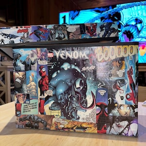 May include: A black wooden box with a collage of comic book images featuring Venom and Spider-Man. The box has the word "VENOM" printed on the front in white letters.