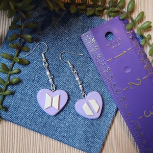 May include: A pair of lavender heart-shaped earrings with white accents and silver hooks. The earrings feature a beaded accent and a logo design. The earrings are displayed on a denim surface.