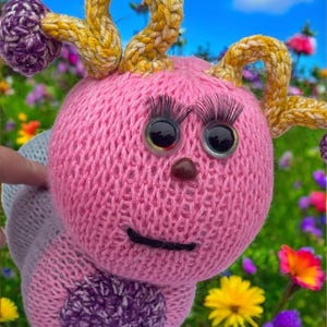 Puede incluir: Un juguete de oruga rosa tejido a mano con grandes ojos saltones, largas pestañas y una pequeña nariz marrón. La oruga tiene antenas amarillas y moradas y un círculo morado en la espalda. El fondo es un campo de flores coloridas.