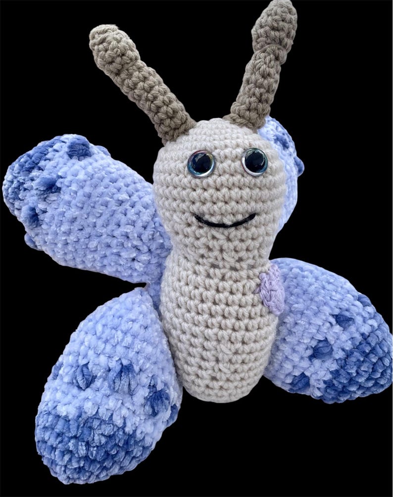 Puede incluir: Un juguete de mariposa hecho a mano con ganchillo, con un cuerpo gris claro, grandes alas azules con manchas azules m&aacute;s oscuras y antenas grises. La mariposa tiene ojos negros grandes y brillantes y una boca sonriente. El juguete est&aacute; hecho de hilo suave.