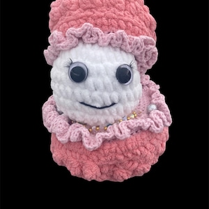 May include: A handmade crochet doll with a white face, large googly eyes, and a smiling mouth. The doll is wearing a pink hat and dress with ruffled edges. A necklace of beads adorns the neck. The doll is set against a black background.