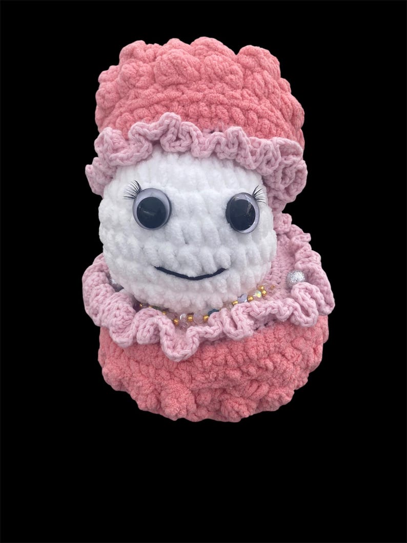 May include: A handmade crochet doll with a white face, large googly eyes, and a smiling mouth. The doll is wearing a pink hat and dress with ruffled edges. A necklace of beads adorns the neck. The doll is set against a black background.