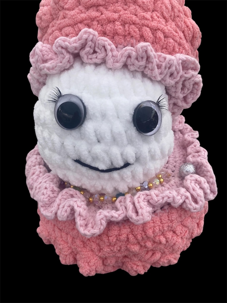 May include: A handmade crochet doll with a white face, large googly eyes, and long eyelashes. The doll is wearing a pink ruffled hat and dress, with a beaded necklace and a silver embellishment. The doll has a smiling expression.