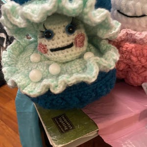 May include: A handmade crochet doll with a mushroom-like design. The doll features a teal and light green frilly hat, white face, blue eyes, and pink cheeks. The doll is sitting on a book that says "Sasquatch Sighting".