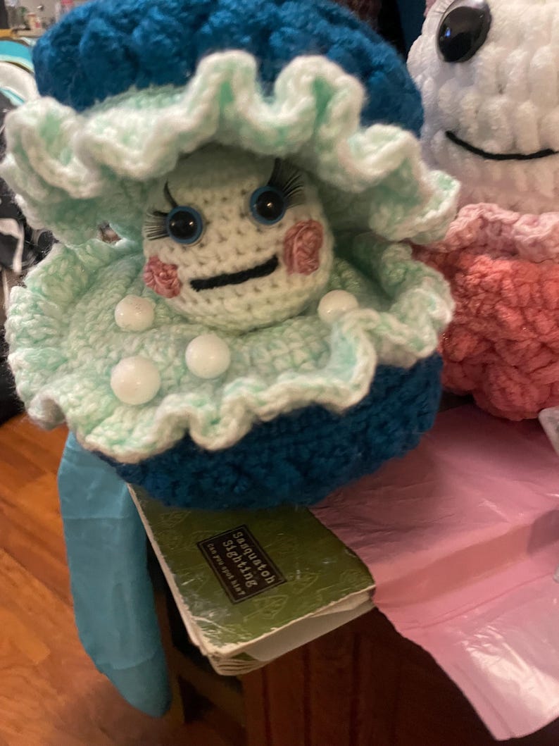 May include: A handmade crochet doll with a mushroom-like design. The doll features a teal and light green frilly hat, white face, blue eyes, and pink cheeks. The doll is sitting on a book that says "Sasquatch Sighting".