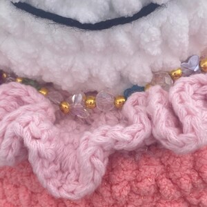 May include: Close-up of a handmade plush toy with a white, fluffy head and a black stitched smile. The toy features a pink, ruffled layer and a beaded necklace with gold and clear beads. The bottom layer is a textured pink.