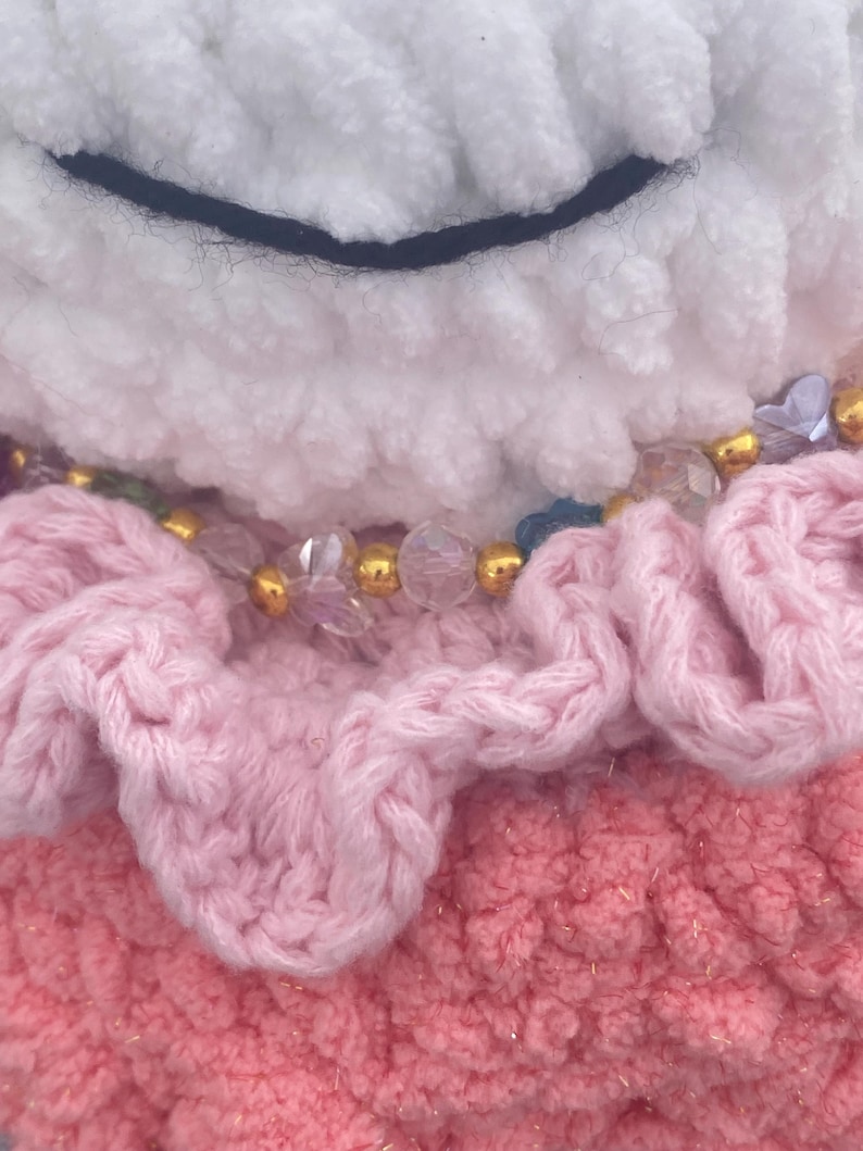May include: Close-up of a handmade plush toy with a white, fluffy head and a black stitched smile. The toy features a pink, ruffled layer and a beaded necklace with gold and clear beads. The bottom layer is a textured pink.