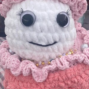 May include: A handmade, plush doll with a white head and a pink dress. The doll has large, googly eyes with eyelashes and a stitched smile. The doll is adorned with a pink ruffled collar and a beaded necklace. The doll's hat is also pink and ruffled.