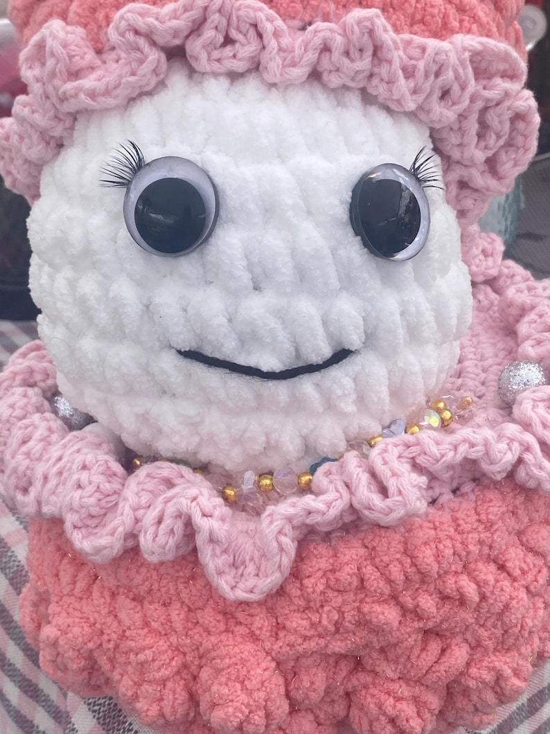 May include: A handmade, plush doll with a white head and a pink dress. The doll has large, googly eyes with eyelashes and a stitched smile. The doll is adorned with a pink ruffled collar and a beaded necklace. The doll's hat is also pink and ruffled.