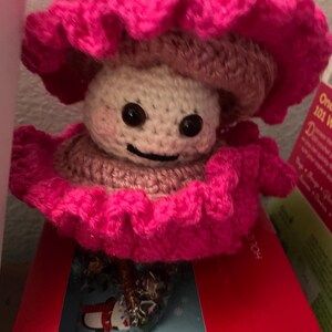 May include: A handmade crochet mushroom doll with a smiling face. The mushroom has a light beige head, a brown cap, and a bright pink ruffled brim. The doll has black button eyes and rosy cheeks. The mushroom is sitting on a red box.