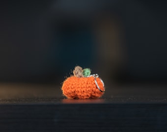 Puede incluir: Un pequeño llavero de calabaza naranja tejido a ganchillo, con un anillo plateado. La calabaza tiene un tallo verde y marrón. El fondo es oscuro y borroso, resaltando los detalles.