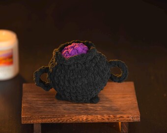 May include: A black crocheted cauldron with two handles, filled with a vibrant purple, textured material. The cauldron sits on a small wooden table. A white candle with a colorful label is in the background.