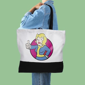 May include: A white canvas tote bag with black handles. The bag features a pink circle with a cartoon woman giving a thumbs up. The woman is wearing a blue jumpsuit and has blonde hair. The text "Vault Girl" is printed on the bag.