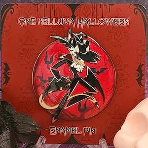 Puede incluir: Un pin de esmalte rojo y negro con un personaje de dibujos animados con un sombrero negro y una capa. El personaje está de pie sobre un fondo rojo con murciélagos. El texto "ONE HELLUVA HALLOWEEN" está en la parte superior del pin y "ENAMEL PIN" en la parte inferior.