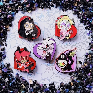 May include: Five enamel pins featuring cartoon characters in heart-shaped frames. The pins display characters in various poses and colours, including red, black, purple, and yellow. The pins are set against a floral background.