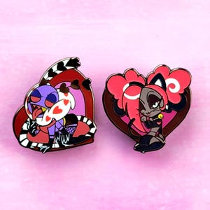 May include: Two enamel pins featuring cartoon characters. One pin shows a purple character with a red and black border, holding a heart-shaped object. The other pin depicts a gray cat with pink hair, set against a heart-shaped background.