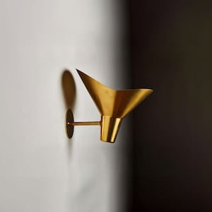 May include: A gold-toned wall sconce with a cone-shaped shade. The sconce is mounted on a white wall.