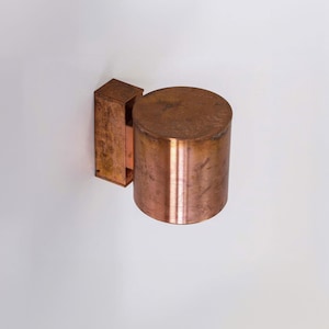 May include: A copper wall sconce with a cylindrical shade. The sconce has a rectangular base and is mounted on a white wall.