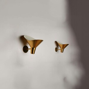 May include: Two gold wall sconces with white interior shades. The sconces are in the shape of a cone and are mounted on a white wall.
